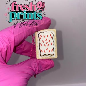 May include: A rectangular pastry-shaped keychain featuring a tan crust, a white frosted center, and small red sprinkles. A pink ball chain is attached to the side of the item. The text "THE fresh prints of Bel-Air" is visible in the upper left corner.