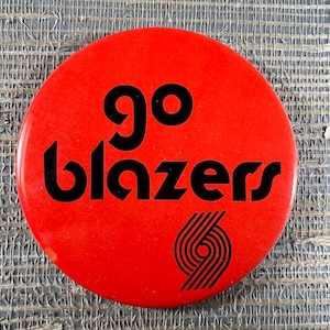 May include: A round, red button with the words "go blazers" in black, along with a black and white logo. The button is a sports-themed item, likely a fan accessory.