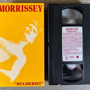 May include: A Morrissey Hulmerist VHS tape and its yellow cardboard sleeve. The sleeve features the name MORRISSEY in red text, a white silhouette of a person, and the title HULMERIST. The black VHS tape has a white label with printed text.