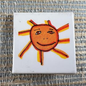 May include: Square white tile featuring a hand-drawn sun design. The sun has an orange face with a smiling expression, brown eyes, and radiating orange and yellow rays. The tile is likely a decorative item.