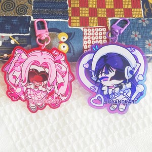 May include: Two acrylic keychains featuring anime-style characters. The left keychain has a pink-haired character with heart-shaped glasses and a pink clasp. The right keychain features a purple-haired character with headphones and a purple clasp. Both have @YANDKART printed on them.