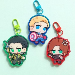 May include: Three acrylic keychains featuring chibi-style characters with colored plastic clasps. One keychain depicts a character with black hair and a green staff, one with blonde hair and a shield, and one with brown hair holding a small black weapon.