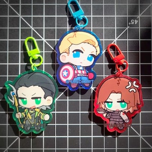 May include: Three acrylic keychains featuring chibi-style characters. One shows a blonde character with a shield, one a dark-haired character with a staff, and one a brown-haired character. Each has a colored clasp and the text @YANDKART printed on them.