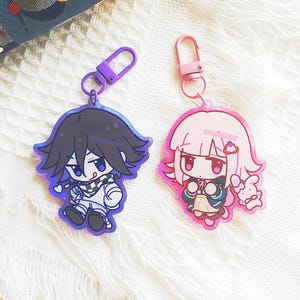 May include: Two acrylic keychains featuring anime-style characters. The left keychain has a purple clasp and a character with black hair and a checkered scarf. The right keychain has a pink clasp and a character with light pink hair and a small rabbit.