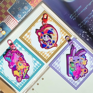 May include: Three acrylic keychains featuring colorful cartoon characters. One shows a jester figure, another a pink geometric creature, and the third a purple rabbit-like character. Each keychain has a matching colored lobster clasp attachment.