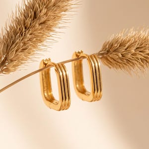 Peut inclure: Une paire de boucles d'oreilles créoles dorées de forme rectangulaire avec trois crêtes parallèles sur la surface extérieure. Les boucles d'oreilles sont placées sur les tiges de deux épis de blé séchés, plumeux et bronzés sur un fond clair.