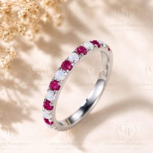 May include: A silver-toned metal ring featuring an alternating row of round-cut, vibrant pink gemstones and iridescent white opals. The stones are set in a shared-prong setting along the top half of the band, creating a continuous, colorful pattern.