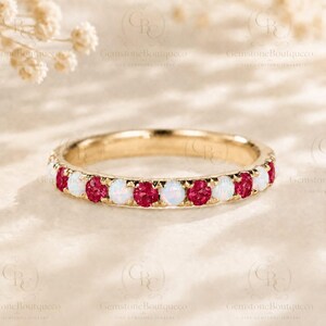 May include: A gold band ring featuring an alternating pattern of round, faceted red rubies and smooth, iridescent white opals set in a continuous row along the outer circumference of the metal band, creating a vibrant and colorful jewelry piece.