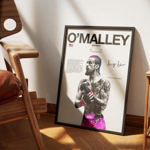 Op de afbeelding: Een ingelijste poster met een zwart-wit portret van UFC-vechter Sean O'Malley met gekleurd haar, handschoenen en een korte broek. De tekst bevat O'MALLEY, SEAN SUGA en een handtekening. Het ontwerp bevat een klein icoon van de Amerikaanse vlag.