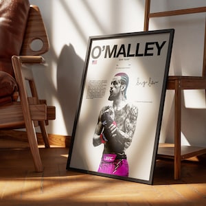 Op de afbeelding: Een ingelijste poster met een zwart-wit portret van UFC-vechter Sean O'Malley met gekleurd haar en een roze vechtbroekje. De tekst luidt O'MALLEY, SEAN "SUGA" en bevat een handtekening op een lichtcr&egrave;mekleurige achtergrond.