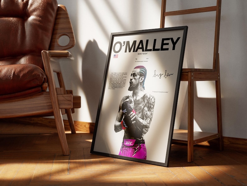 Op de afbeelding: Een ingelijste poster met een zwart-wit portret van UFC-vechter Sean O'Malley met gekleurd haar en een roze vechtbroekje. De tekst luidt O'MALLEY, SEAN "SUGA" en bevat een handtekening op een lichtcr&egrave;mekleurige achtergrond.