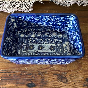 May include: A rectangular ceramic baking dish featuring a deep blue and white floral pattern. The interior and exterior are decorated with repeating circular motifs and small star-like shapes in the center, finished with a solid blue rim.