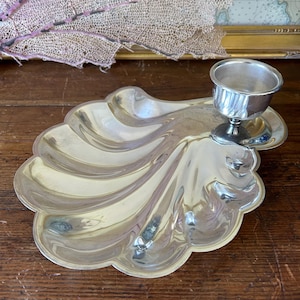 May include: A polished silver-tone metal tray shaped like a large scallop shell, featuring a smooth, reflective surface with deep, radiating ridges. A small, cylindrical candle holder cup is attached to the upper right side of the shell tray.