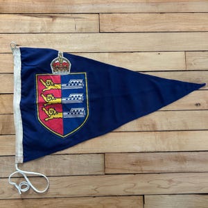 May include: A triangular navy blue pennant flag featuring a central shield emblem divided into red and blue sections. The red side displays three yellow lions, while the blue side shows three white horizontal bars. A crown sits above the shield.