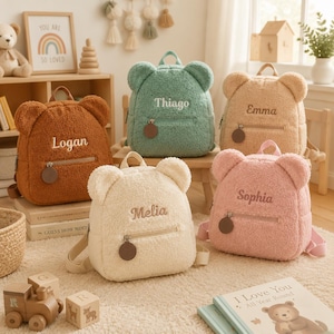 Può includere: Cinque zainetti in peluche a forma di orso nei colori marrone, verde salvia, beige, crema e rosa. Ogni zaino presenta una tasca frontale con cerniera e tirante circolare, orecchie da orso sulla parte superiore e un nome ricamato sul davanti.