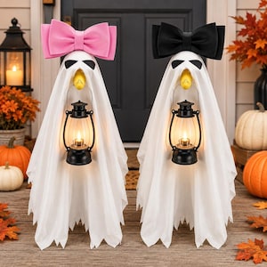 May include: Two ghost decorations draped in white fabric, each featuring a yellow beak, black eye markings, and a black lantern. One ghost wears a large pink bow, while the other wears a large black bow. Both figures stand upright on a wooden surface.