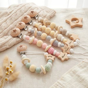 May include: Five dummy clips featuring wooden star-engraved clips, silicone and crochet beads in pastel colours, and wooden star charms engraved with the names Noah, Rosie, Waylon, and Julius, arranged on a textured fabric surface.