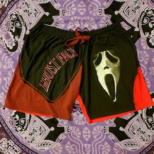 May include: Black shorts with a red drawstring waist, featuring the text GHOST FACE in red on the left leg and a white Ghost Face mask graphic on the right leg. The shorts have red side panels and a black base with a red interior lining.