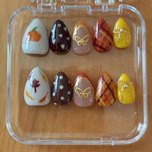 Könnte beinhalten: Ein Set aus zehn mandelförmigen Press-on-Nägeln in einem durchsichtigen Kunststoffetui. Die Designs umfassen weiße Nägel mit Kürbissen und Blättern, dunkelbraune Nägel mit Herzen und Punkten, hautfarbene Nägel mit goldenen Schmetterlingen, Karomuster und gelbe Nägel mit goldenen Schleifen.