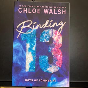 May include: A paperback book titled Binding 13 by Chloe Walsh, part of the Boys of Tommen series. The cover features a dark blue background with a smoky effect and the number 13 rendered in a sparkling, multi-colored glitter texture.