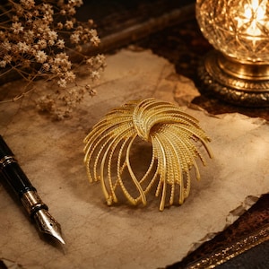 May include: A gold-toned brooch featuring a circular, fan-like arrangement of textured, radiating wire strands that curve outward from a central point, creating a layered, dimensional appearance with a polished metallic finish.