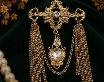May include: A gold-toned metal brooch featuring an ornate, symmetrical design with clear, faceted crystals. The piece includes two long, dangling gold chain tassels on either side and a central pendant element suspended by a delicate chain.
