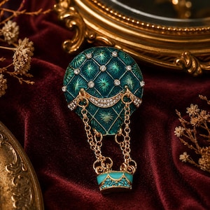 May include: A brooch shaped like a hot air balloon with a teal quilted enamel canopy featuring embedded clear crystals. The balloon is connected by gold-toned metal chains to a small teal basket accented with a row of small blue crystals.