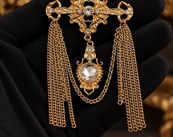 May include: A gold-tone metal brooch featuring an ornate cross-shaped top with clear rhinestones and a central circular stone. Two long, multi-strand gold chains hang from the sides, connected by a central dangling pendant with a clear stone.