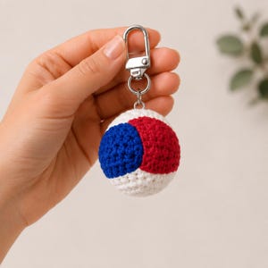 May include: A spherical crochet keychain featuring a color-blocked design in white, royal blue, and red yarn. The round textile charm is attached to a silver-toned metal lobster clasp and a circular split ring, providing a secure attachment point.