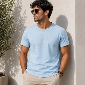 May include: A light blue short-sleeved t-shirt with a crew neckline and a subtle heathered texture. The garment is worn with beige trousers, dark-tinted sunglasses, and a wristwatch, presenting a simple and clean aesthetic for casual wear.