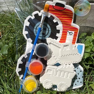 May include: A craft kit in clear plastic packaging containing a blue paintbrush, three small circular paint pots in black, orange, and yellow, and two white textured construction vehicle shapes. A red and yellow tractor cutout is included.