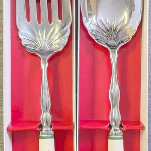 Puede incluir: Un juego de tenedor y cuchara para servir en tono plateado con cuencos de metal pulido que presentan un diseño en forma de concha en la base. Los mangos son de cerámica blanca, decorados con una ilustración de un árbol de Navidad colorido.