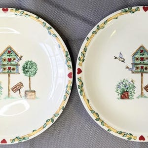 May include: Two round cream-colored ceramic plates featuring a central illustration of a teal birdhouse on a post, a small topiary, and a flowering plant. The rim is decorated with a yellow ribbon pattern, green vines, and small red hearts.