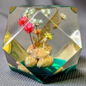 Puede incluir: Un pisapapeles geométrico de resina transparente y facetada que contiene un grupo de pequeñas flores secas en tonos rojo, blanco y amarillo, junto con varias conchas marinas de color tostado y moteado incrustadas en la base.