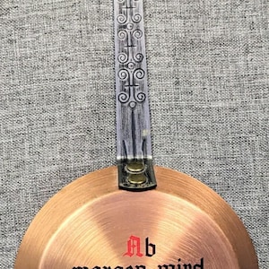 May include: A round copper wall plaque with a long metal handle featuring an etched scroll pattern. The center displays the black German text "Ab morgen wird gespart" above a colorful floral design. Three brass hooks are attached to the bottom.