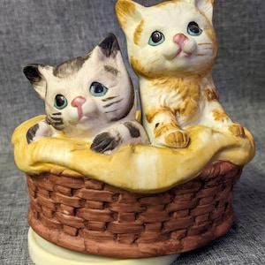 Puede incluir: Una figura de cerámica con dos gatitos sentados dentro de una cesta tejida marrón. Un gatito es naranja y blanco, mientras que el otro es gris, negro y blanco. Ambos gatitos tienen ojos azules y nariz rosa, sobre una tela amarilla en la cesta.