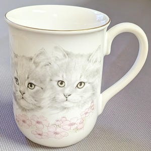 May include: A white ceramic mug featuring a detailed illustration of two fluffy gray kittens side by side. The base of the mug is decorated with a delicate pattern of small, light pink flowers. The rim of the mug is finished with a thin gold trim.