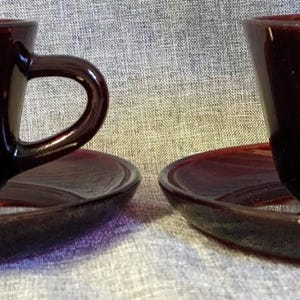 May include: Two matching dark burgundy glass teacups with rounded bowls and curved handles, each resting on a coordinating circular glass saucer. The glassware features a smooth, glossy finish with a deep red translucent tone throughout the set.