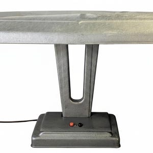 May include: A silver-toned metal desk lamp featuring a wide, rectangular shade and a V-shaped support column. The rectangular base includes two small circular buttons, one red and one black, with a thin black power cord extending from the side.