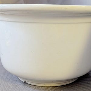 May include: A solid white ceramic chamber pot featuring a smooth, glossy finish, a wide circular rim, and a single curved handle on the side. The vessel has a deep, rounded basin and a sturdy base, presenting a clean, uniform appearance throughout.