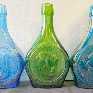 May include: Three glass bottles with iridescent finishes, each featuring a circular embossed portrait of a military figure. The left bottle is light blue, the centre is lime green, and the right is a deep teal, all with narrow necks and wide bases.