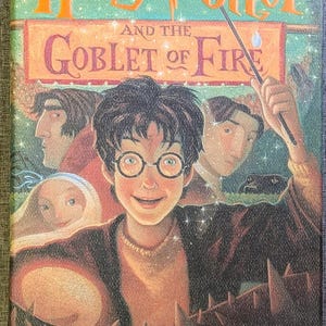 May include: The hardback book Harry Potter and the Goblet of Fire by J.K. Rowling features a colourful illustration of Harry Potter with glasses, surrounded by other characters, a wand, and a glowing light against a dark green background.