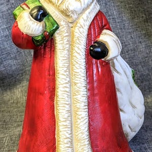 May include: A ceramic Santa Claus figurine wearing a red coat with white fur trim and a matching hat. The figure has a long white beard, holds a green gift box in one hand, and carries a large white sack over the other shoulder. The surface has a textured finish.