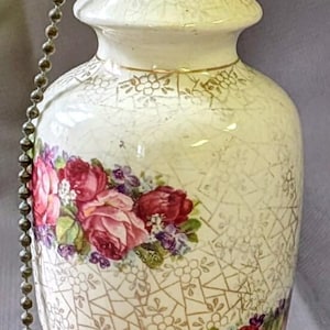 May include: A ceramic lamp base featuring a cream-colored body with a gold-toned geometric lattice pattern and floral decals of pink roses and purple flowers. The base has a crackled glaze finish and a metal socket with a pull chain switch.
