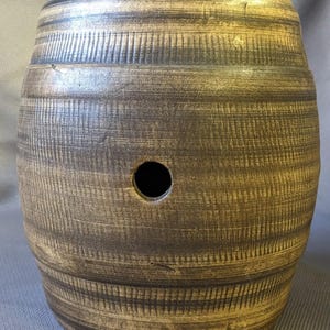 May include: A barrel-shaped ceramic vessel with a textured, weathered brown finish featuring dark horizontal streaks and a single circular opening centered on the front. The surface displays a wood-grain appearance with subtle vertical etched lines.