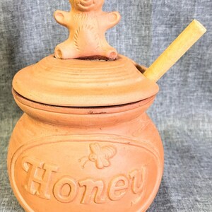 May include: A terracotta honey pot with a lid featuring a sculpted bear figure on top. The pot has the word Honey embossed on the front alongside a small bee icon. A light-colored wooden honey dipper is inserted through a notch in the lid.