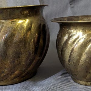 Puede incluir: Dos maceteros de latón con forma redondeada y bulbosa y un distintivo patrón estriado en espiral. Las superficies metálicas presentan un acabado dorado pulido con pátina natural y signos de antigüedad, con bordes anchos y acampanados.