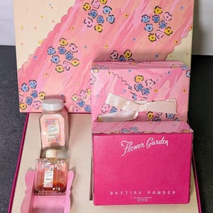 May include: A Flower Garden gift set featuring a sachet, a perfume bottle, and a box of dusting powder. The packaging is pink with a floral pattern and white ribbon accents. The dusting powder box is bright pink with white text reading Flower Garden.