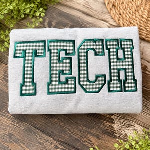 Puede incluir: Una sudadera gris claro con la palabra TECH en letras mayúsculas grandes. Las letras están rellenas con un patrón de cuadros vichy verde y blanco y están delineadas con un borde grueso bordado en verde oscuro sobre la tela.