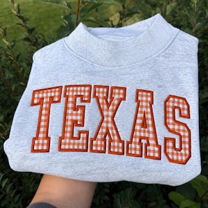 Puede incluir: Una sudadera gris claro de cuello redondo con la palabra TEXAS bordada en el pecho en una fuente universitaria. Las letras están rellenas con un patrón de cuadros vichy naranja y blanco y delineadas con un borde de hilo naranja sólido.